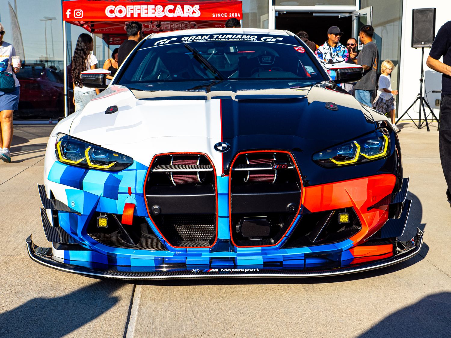 BMW M4 in a blue, red and white Gran Turismo livery, head-on under the Coffee and Cars tent